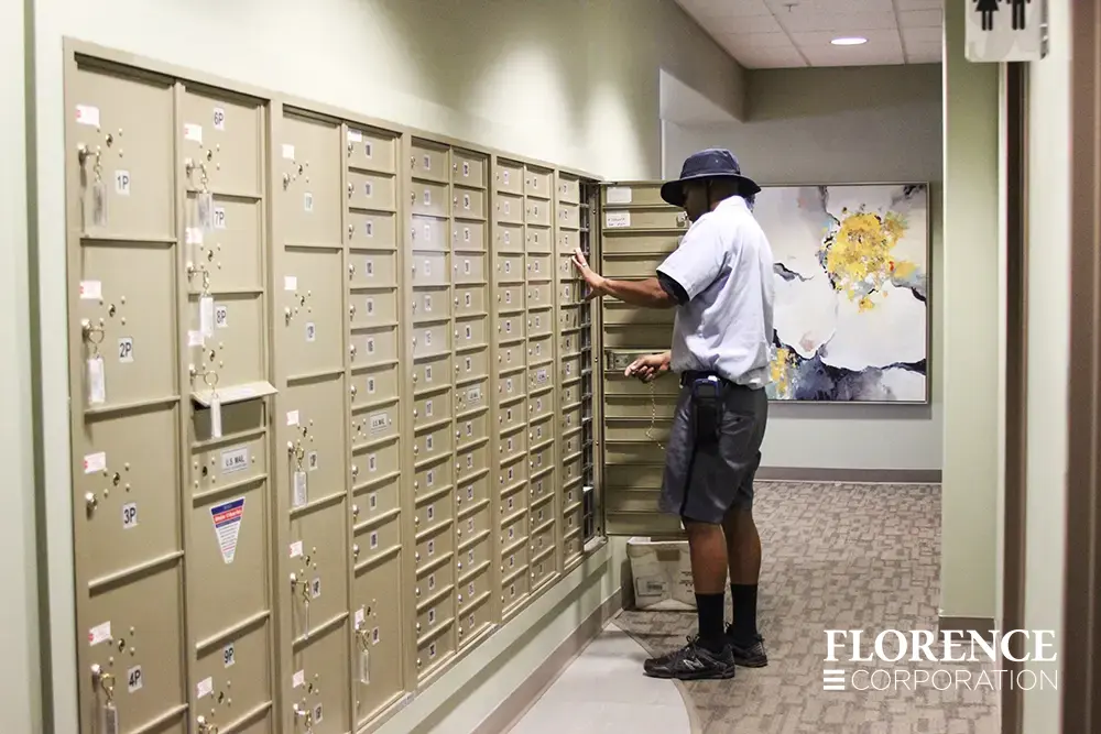 Recessed mount versatile™ 4C mailboxes and parcel lockers in sandstone installed in a hallway of a modern multifamily apartment building with floral painting in background and usps postal carrier delivering mail with master access door open