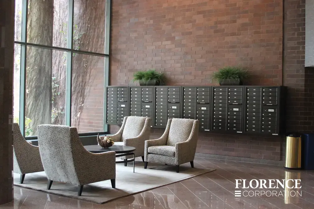 surface mounted versatile™ 4C mailboxes in dark bronze mounted with matching collar indoor on red brick wall near four chairs and a table